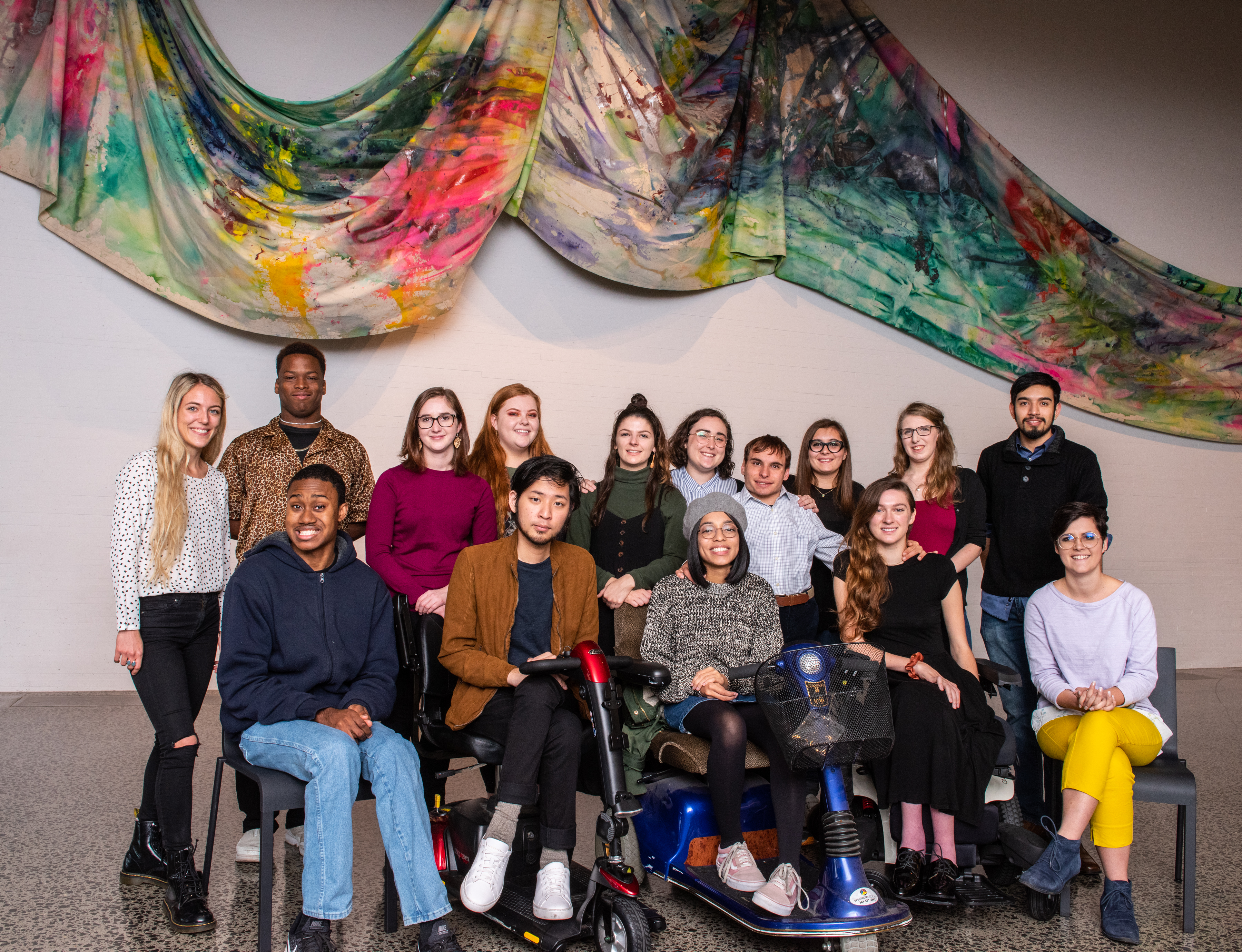 A photo of the 15 winners of the 2019-2020 Emerging Young Artists Program, posed in two rows, the front row seated, under a multicolored swirling horizontal sculpture