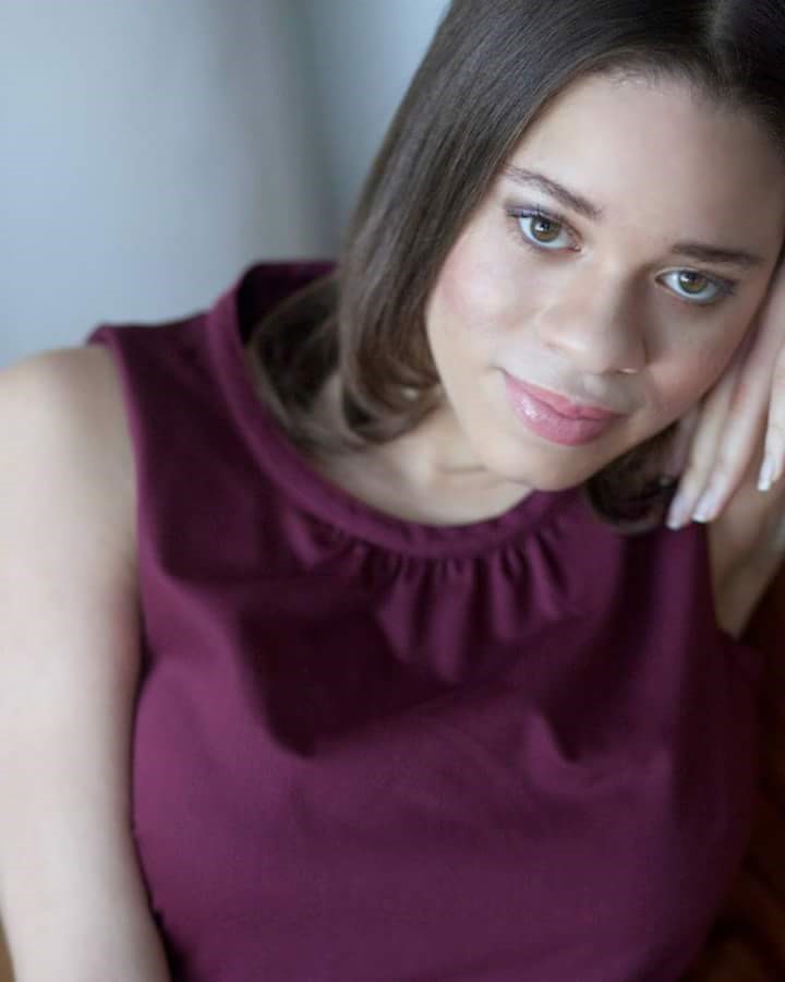 [Image Description:  A woman with dark hair and brown eyes.  Her face is in the upper right corner of the photo.  She is leaning against her hand and wearing a purple shirt.]