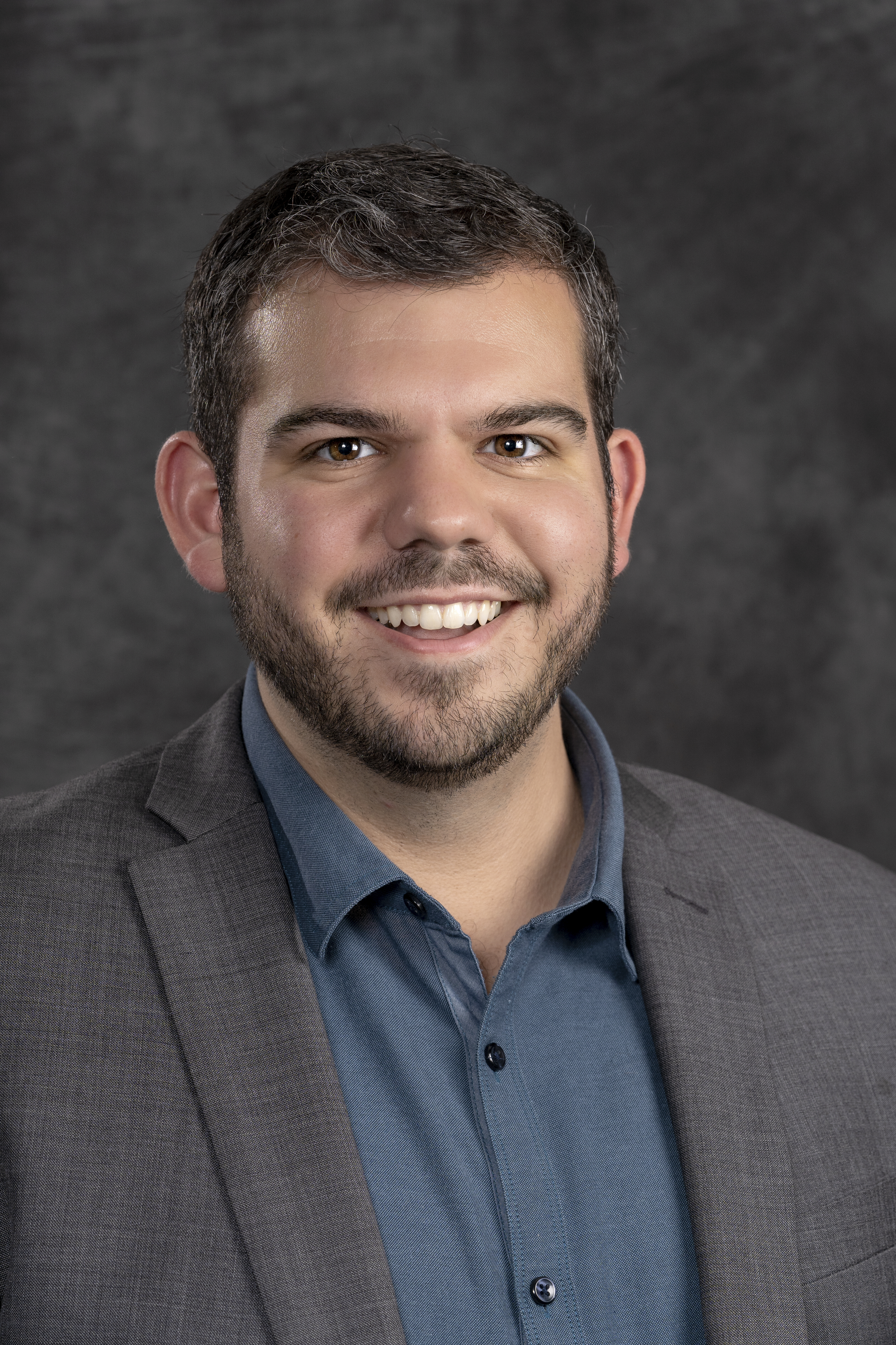 Image Description: A headshot of a man with brown hair and a beard.  He is smiling, wearing a blue shirt and brown coat, in front of a neutral background.]