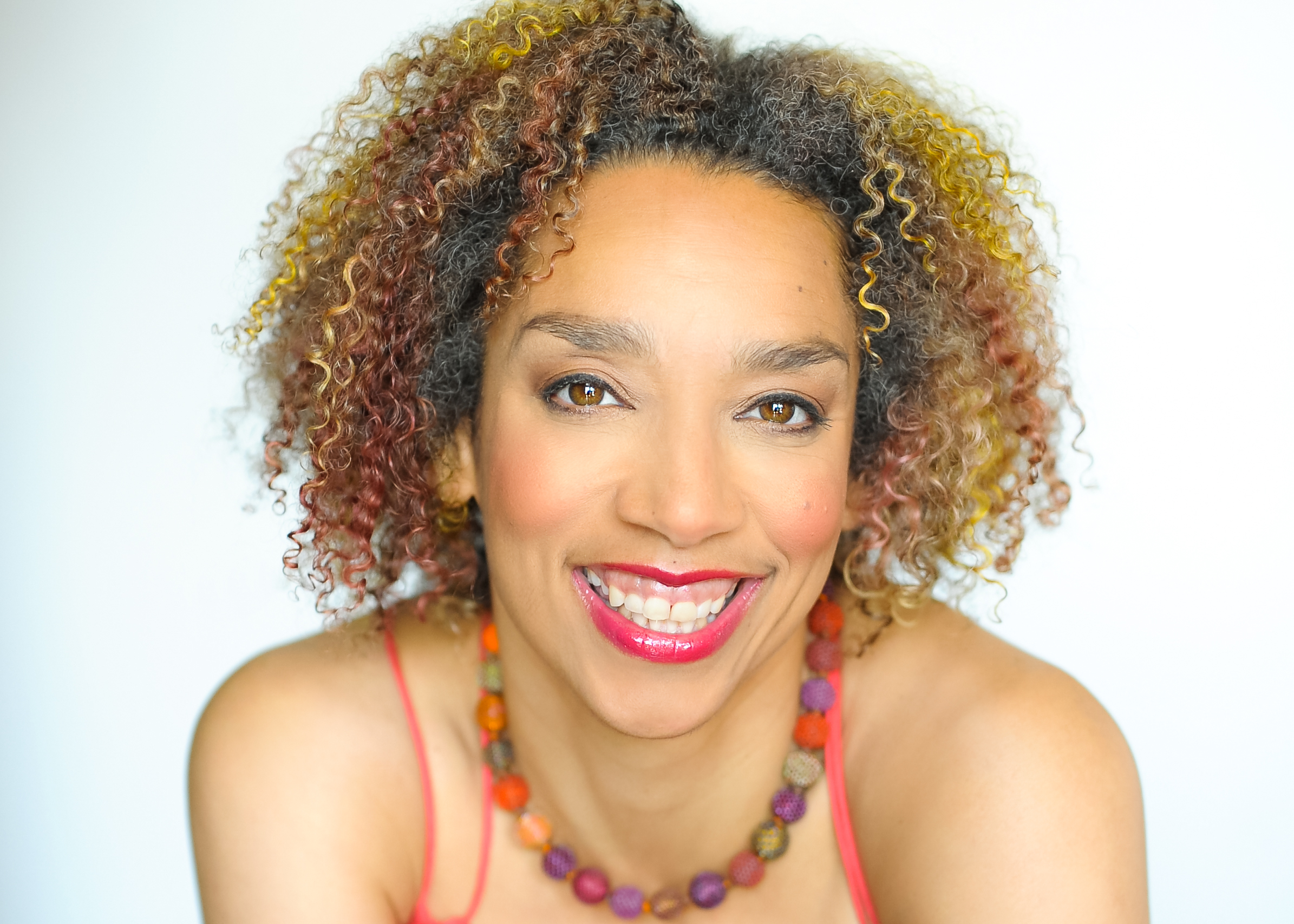 Alice is a light skinned multi-racial woman with brown, yellow and copper streaks in her curly hair. She smiles and gazes at the camera. A necklace of Autumn colored beads sits around her neck. Photo by Beverlie Lord