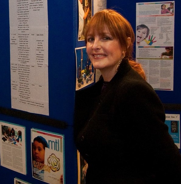 [Image Description:  A photo of a smiling woman with red hair, dressed in black.  She is standing in front of a blue tri-fold board with photos and text related to children and visual art.]