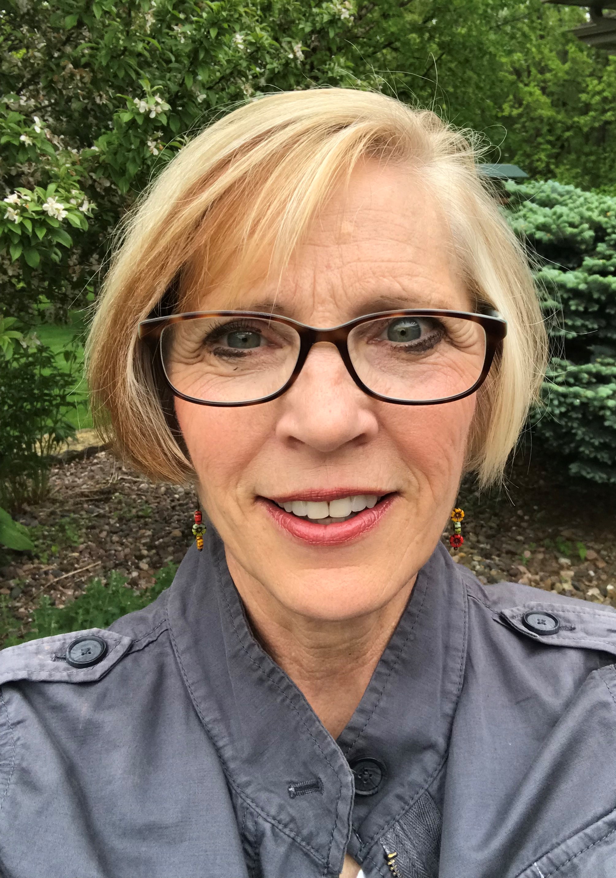 [Image Description: A photo of a woman with short blonde hair and brown glasses.  Her face is central in the image, against an outdoor background with trees.]