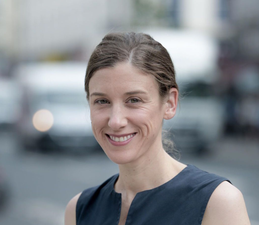 A smiling woman with brown hair.  She is in front of a blurred urban background.