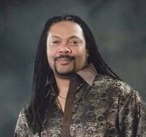 A headshot of a man with long brown hair and a goatee.  His face is central in the picture and he wears a brown patterned shirt.