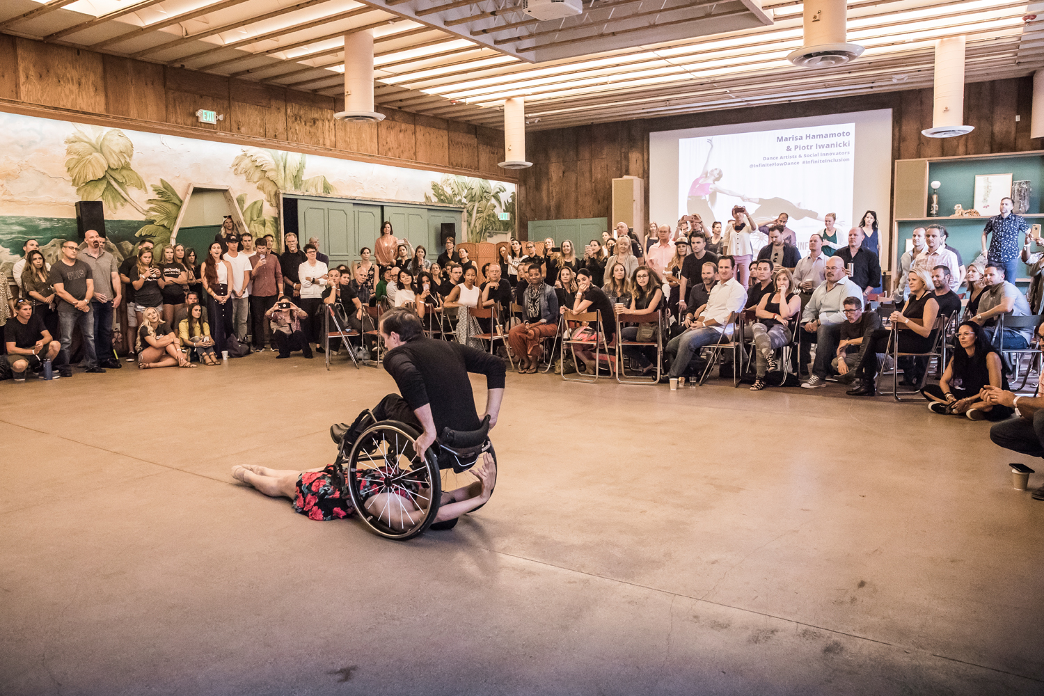 Image: One dancer lies beneath another in a wheelchair. We see the back of the dancer in the wheelchair. An audience watches them in a semicircle, some people seated and some standing. The audience and dancers are on the same level; there is no stage.