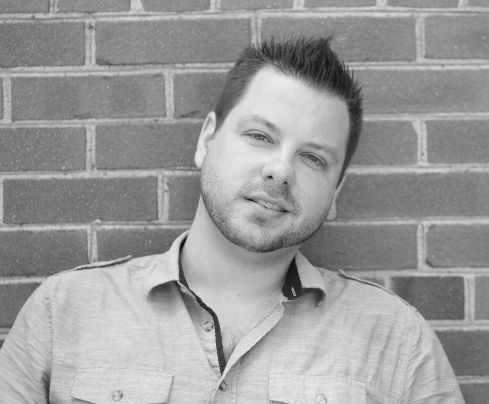 A black and white headshot of a man with short hair.  He is leaning against a brick wall.