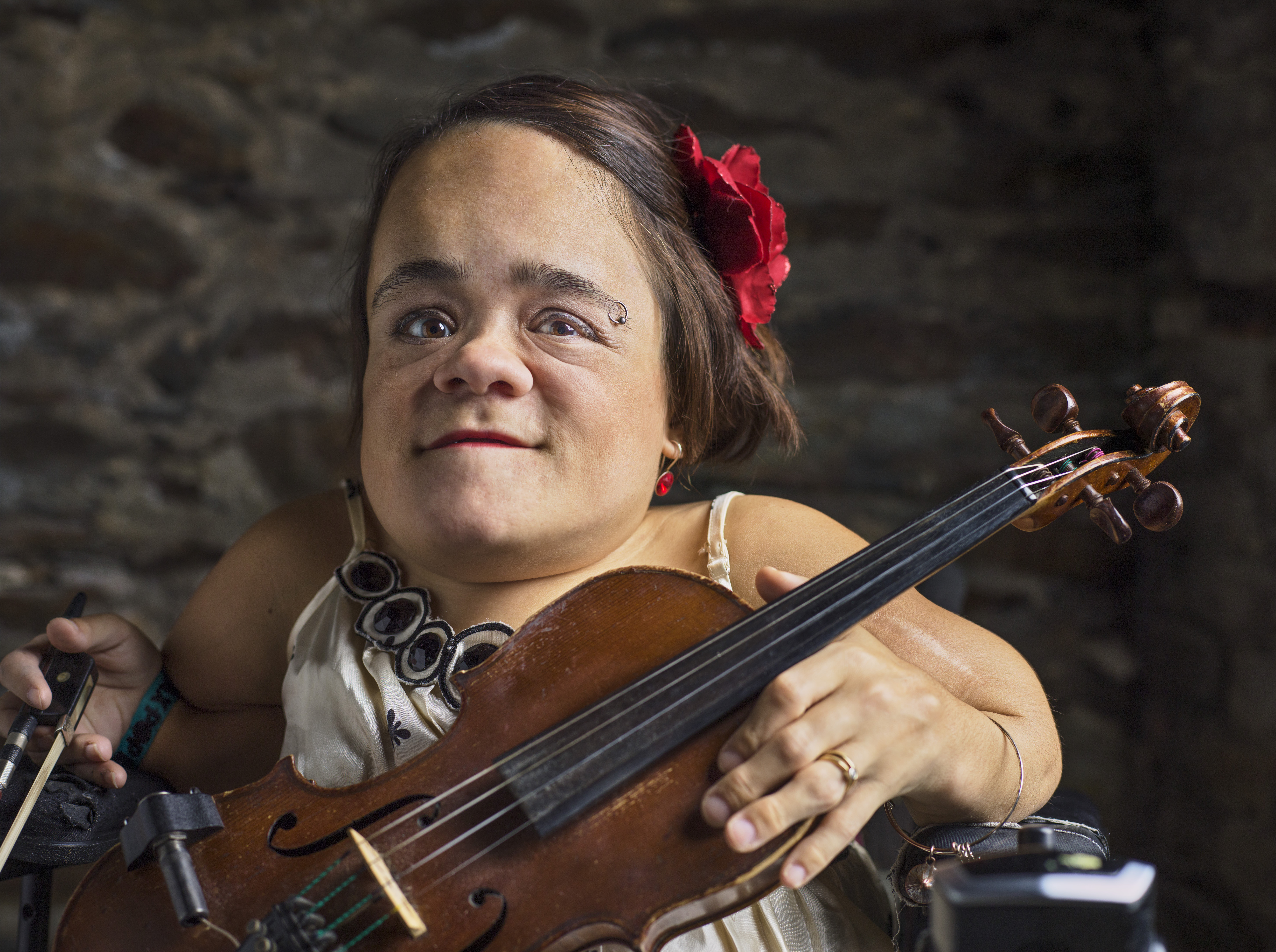 Photo of Gaelynn Lea, a woman holding a violin in her left hand and a bow in her right hand. Gaelynn is wearing a white sleeveless shirt and red flower in her chin-length brown hair. Her left elbow rests on the arm of her wheelchair. Photo by Paul Vienneau, paulvienneau.com