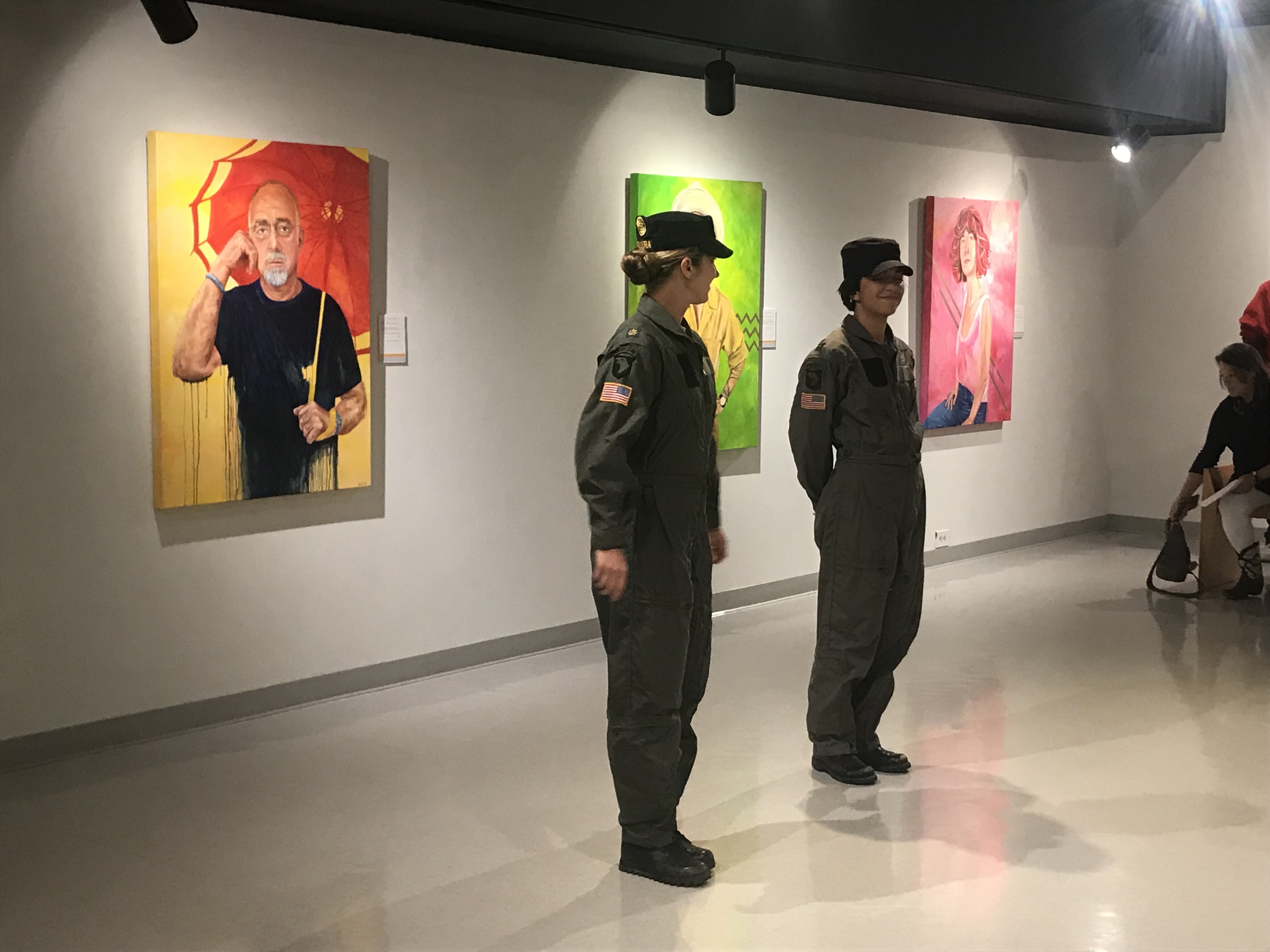 Two women in army dress standing in front of three colorful portraits. The portrait on the left of a man with a white bear holding a red parasol against a yellow background, the middle portrait is partially hidden behind the two women, and the far right portrait is of a person with red hair in front of a pink background. 