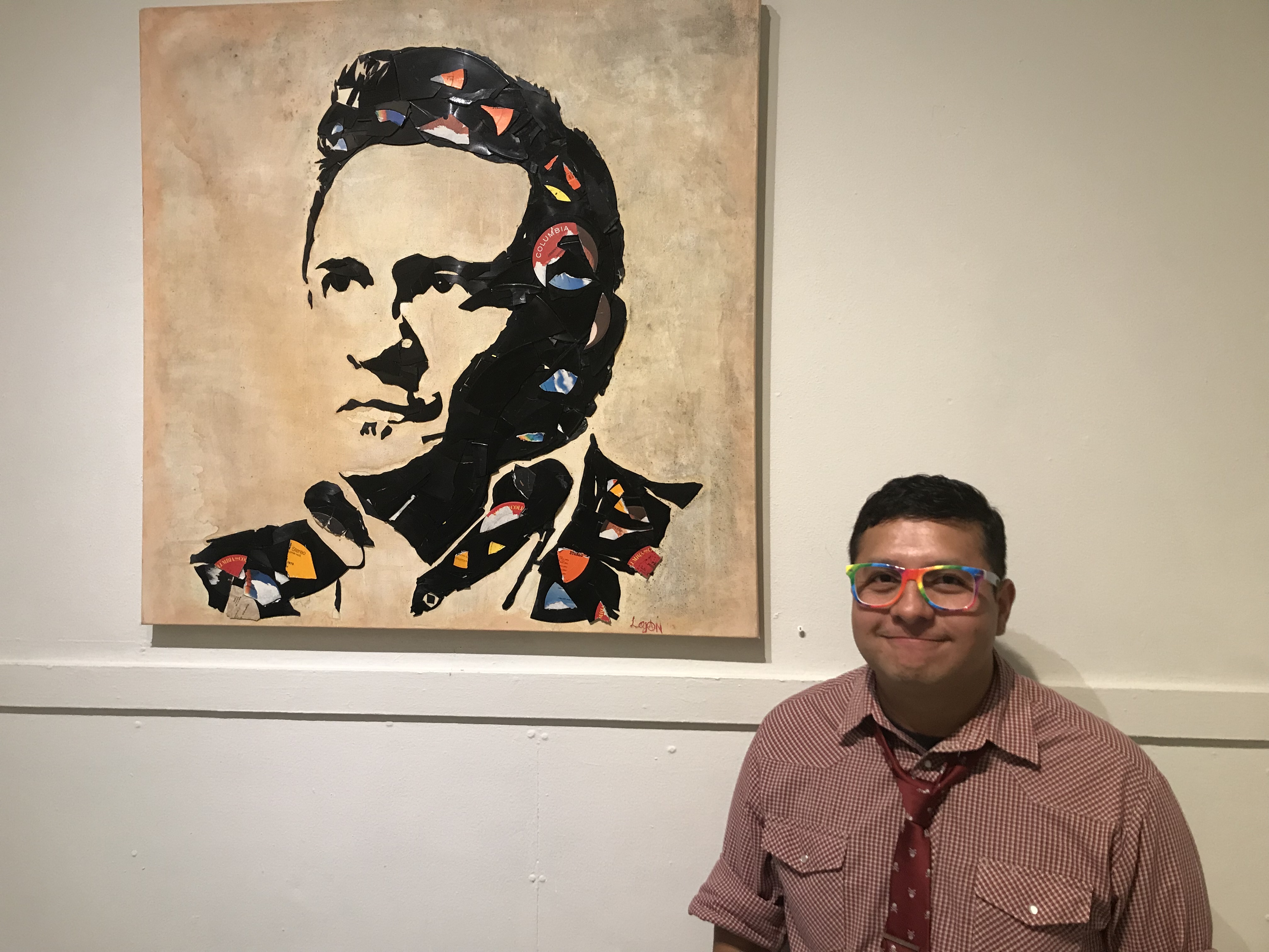 A man with short dark hair wearing a red shirt and rainbow glasses stands in front of a portrait of a man