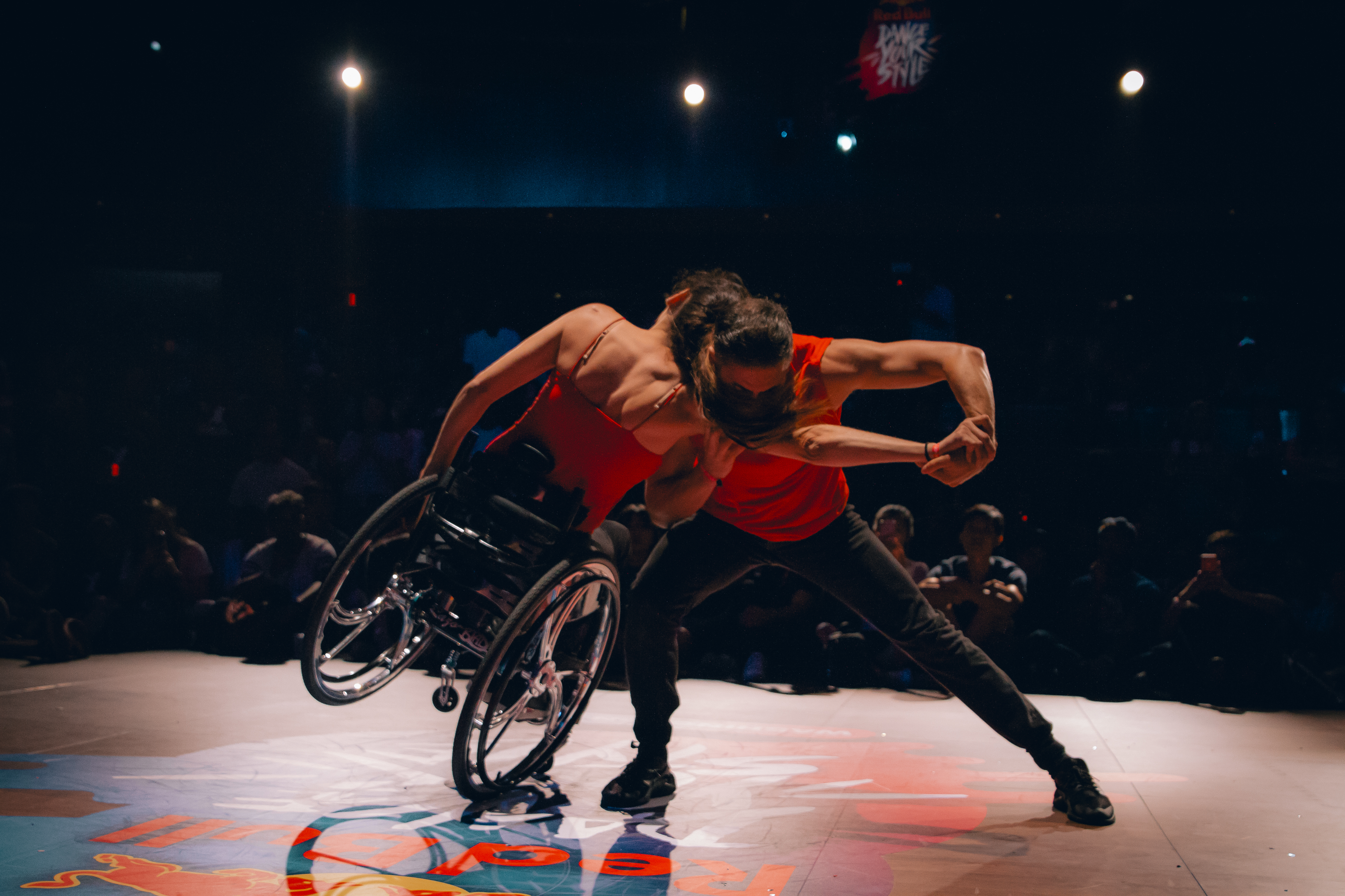 Image: a dancer in a wheelchair is shown from behind, leaning into a standing dancer. The standing dancer's arm is outstretched towards their partner.Audiences members watch in a semicircle, on the same level as the dancers. There is no elevated stage.