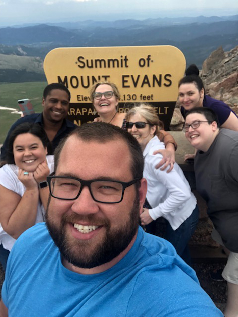 Image: a selfie of group of seven adults smiling in front of a sign that reads 