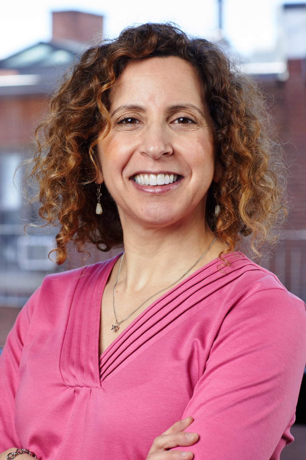 Headshot of woman with brown curly hair wearing a pink shirt.  She is smiling at the camera in front of a background of brick buildings.