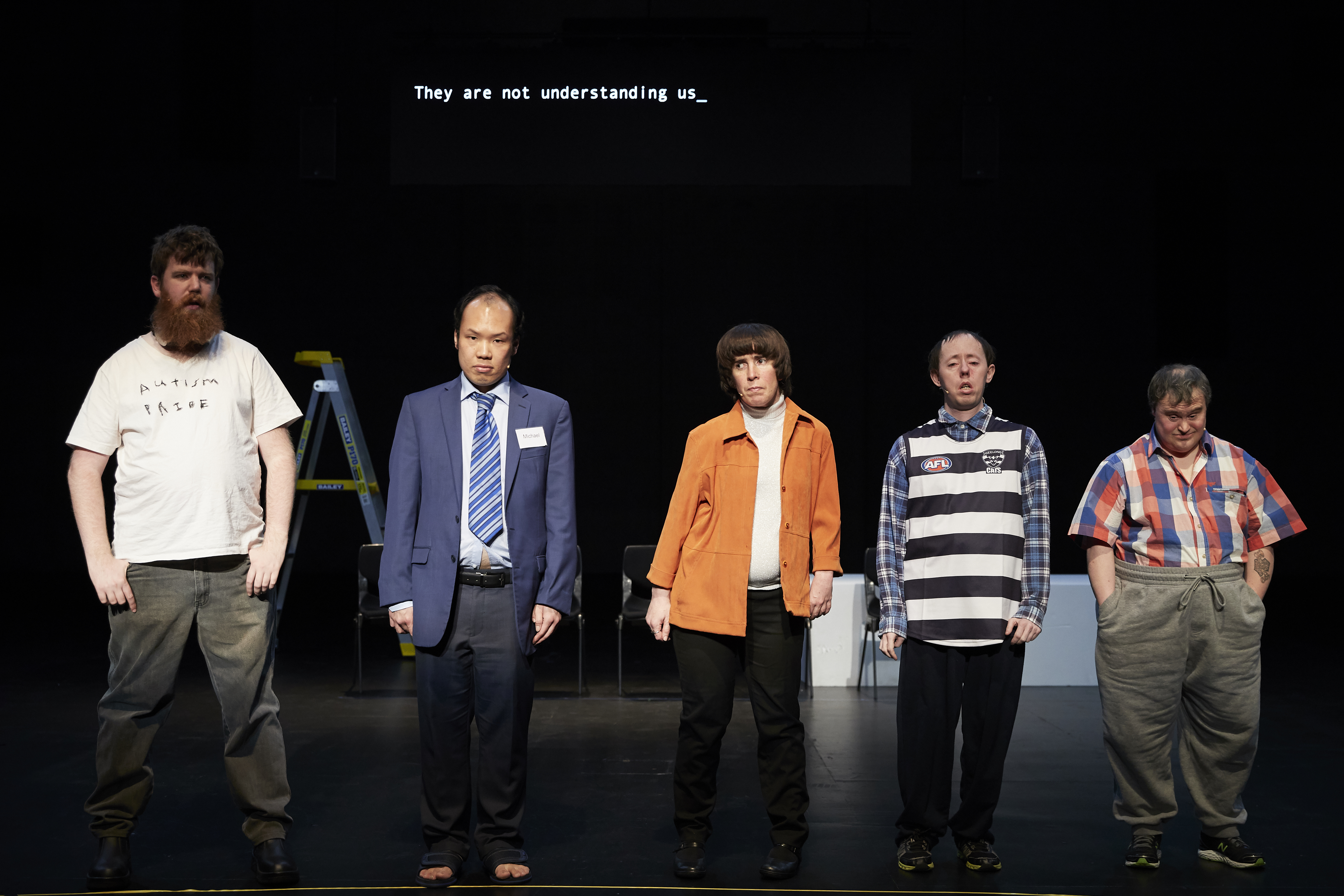 Image from the show THE SHADOW WHOSE PREY THE HUNTER BECOMES, of five actors on a stage facing the audience; the tallest actor is on the left, and they are in descending height order with the shortest on the right. Above the actors heads are the projected words, 