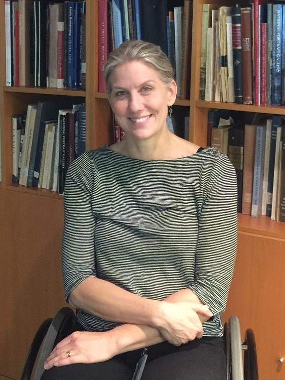 Blonde woman sitting and smiling at the camera, her arms are crossed in her lap