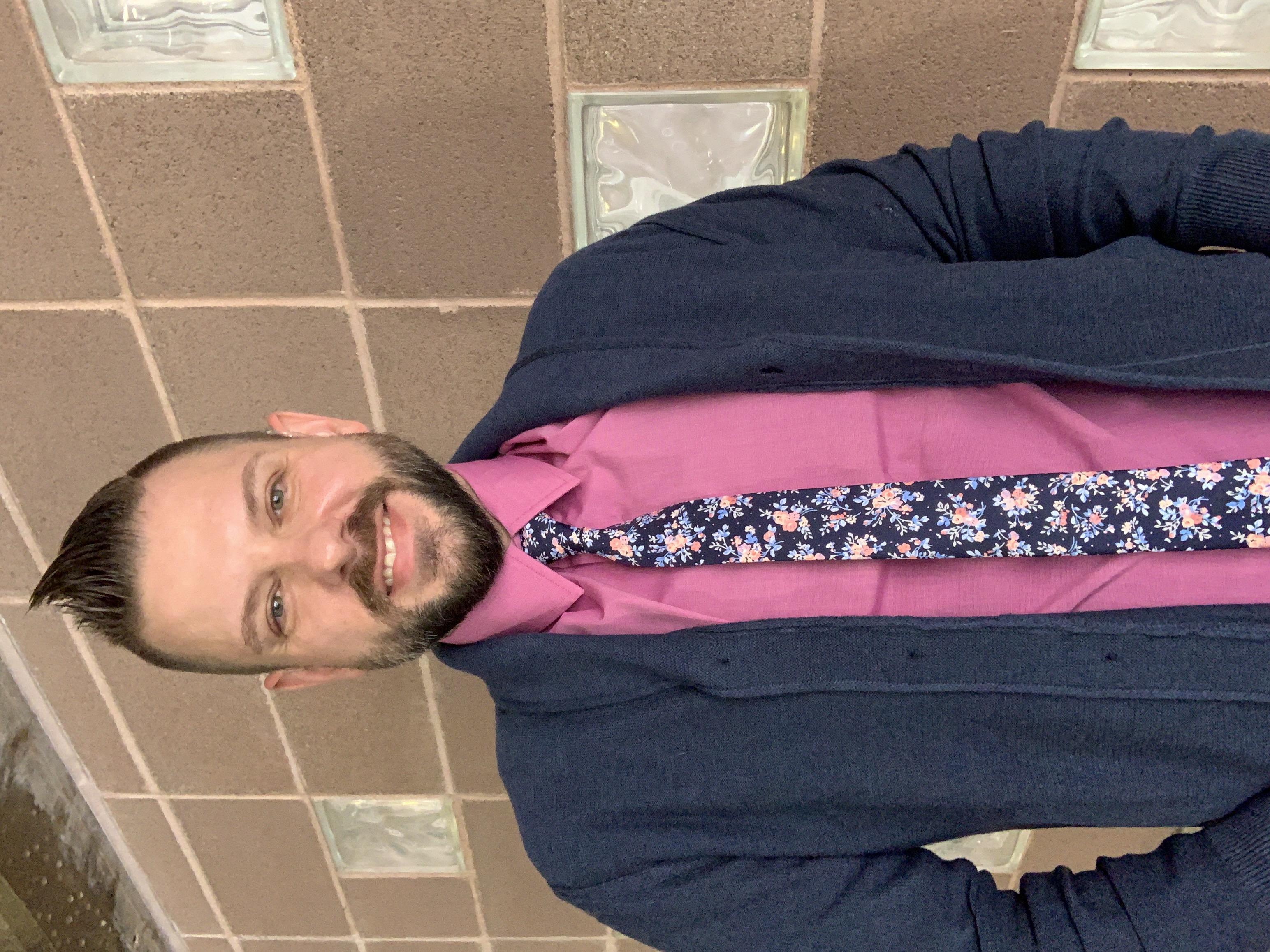 Smiling man with short brown hair and beard, wearing pink button down shirt, dark blue cardigan sweater, blue floral tie.