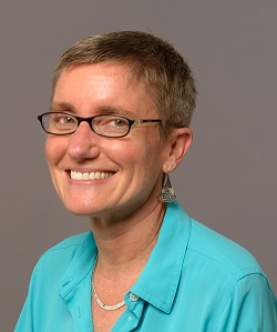 A smiling worman with very short straight blond hair and light colored skin wearing dark framed glasses and a light blue button down blouse.