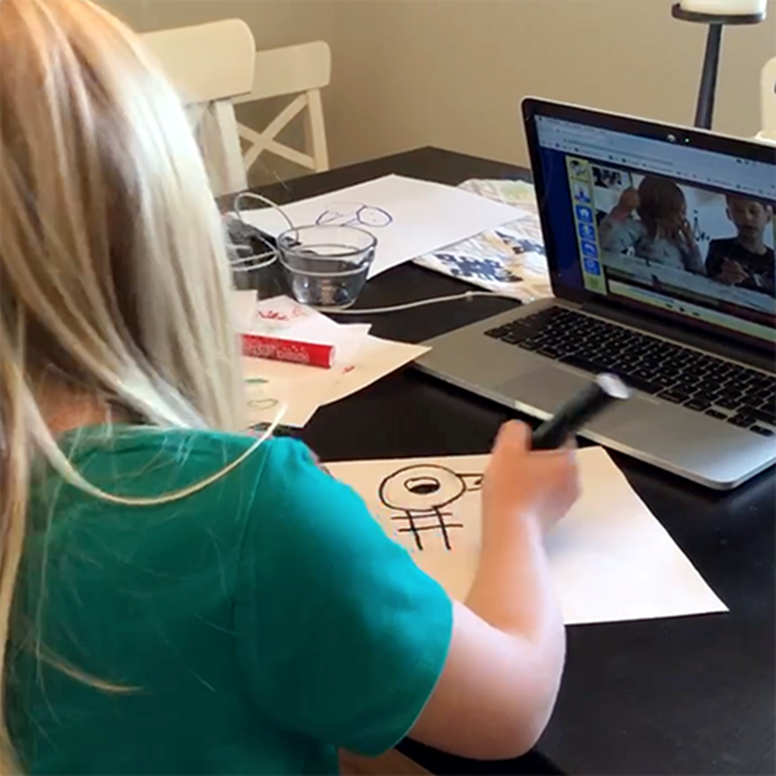 A child with blonde hair and a green shirt sits, facing away from the camera, in front of a computer.  She is drawing Pigeon from the Mo Willems series on white paper with a black marker.
