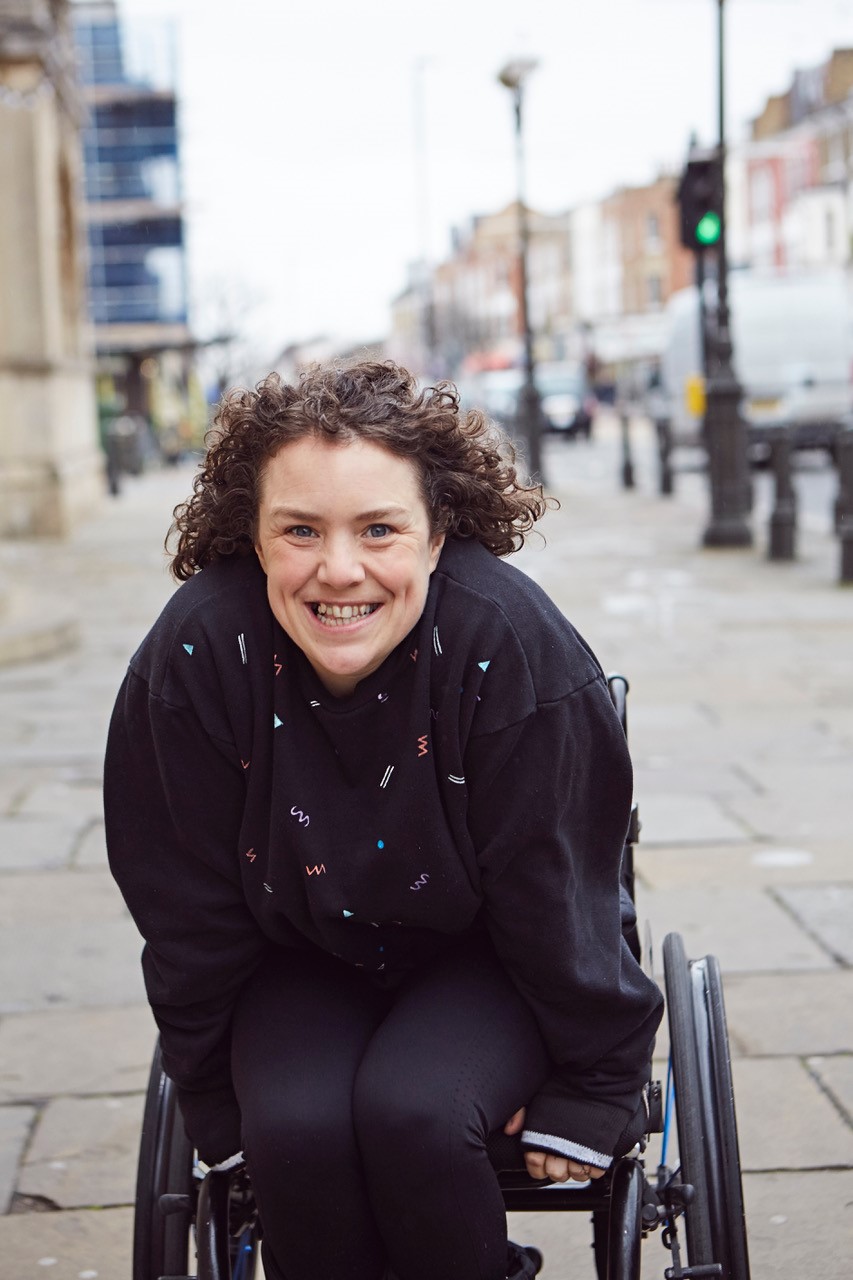Photo by James Lyndsay. Photo of Jessica Thom, a white woman with curly brown hair in a wheelchair, wearing a black shirt and smiling; she is on a street in daytime.