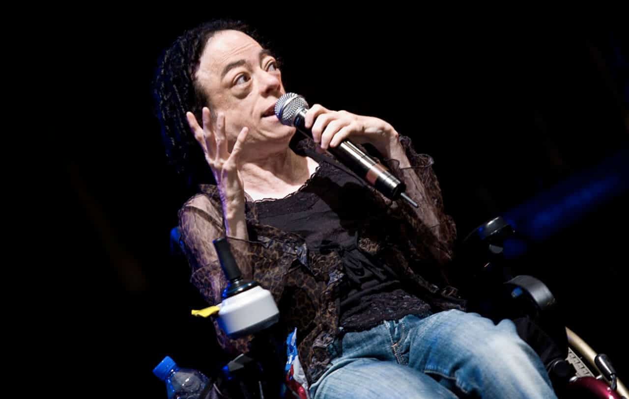 Photo of Liz Carr, a white woman with dark, short, curly hair in a wheelchair, holding a microphone with her left hand. She is wearing a black top with lace trim and blue jeans.